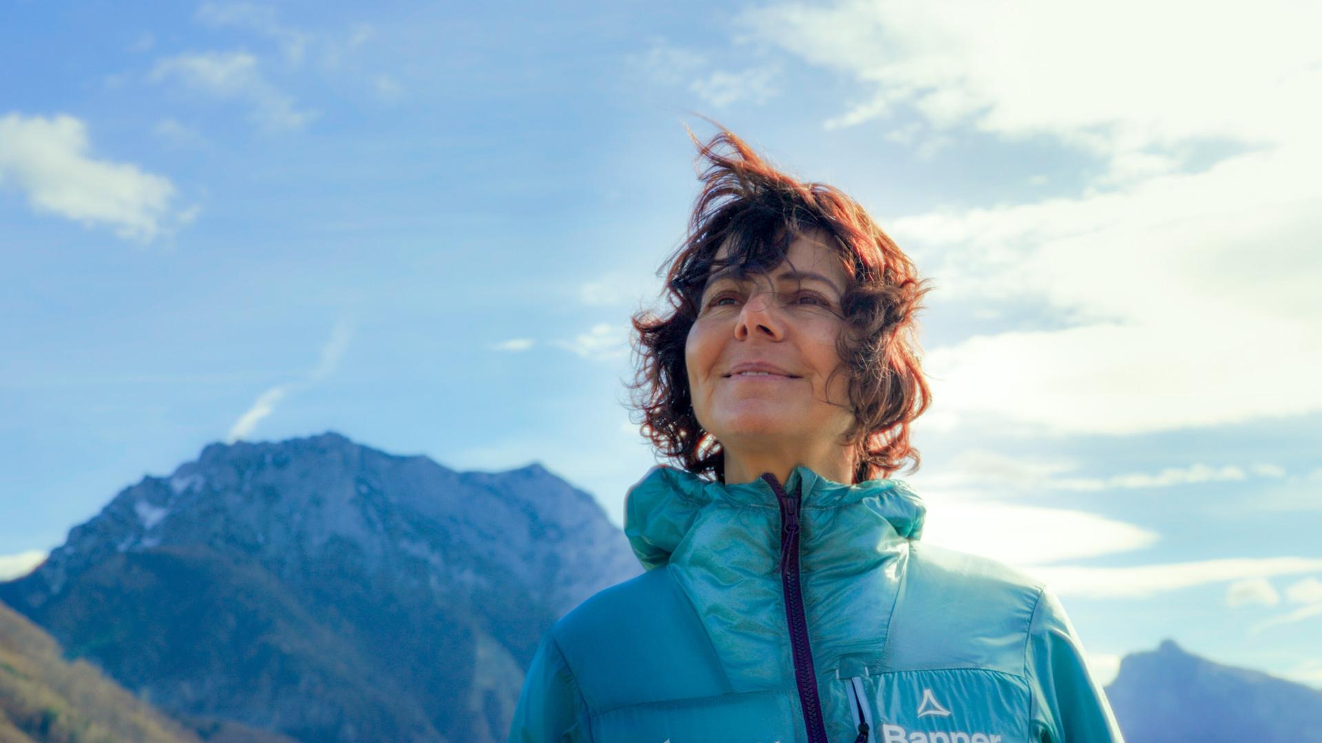 Das Bild zeigt eine Frau, die im Freien vor einer beeindruckenden Berglandschaft steht. Sie hat lockiges, braunes Haar, das vom Wind aufgewirbelt wird. Die Frau trägt eine grüne Jacke, die warm und funktional aussieht, und lächelt leicht, während sie in die Ferne blickt. Im Hintergrund sind große, felsige Berge zu sehen, die teilweise mit Schnee bedeckt sind. Der Himmel ist teilweise bewölkt mit einigen kleinen, weißen Wolken und hat eine insgesamt helle, freundliche Atmosphäre. Die Umgebung wirkt natürlich und unberührt, was auf eine alpine Landschaft hindeutet.