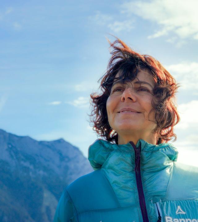 Das Bild zeigt eine Frau, die im Freien vor einer beeindruckenden Berglandschaft steht. Sie hat lockiges, braunes Haar, das vom Wind aufgewirbelt wird. Die Frau trägt eine grüne Jacke, die warm und funktional aussieht, und lächelt leicht, während sie in die Ferne blickt. Im Hintergrund sind große, felsige Berge zu sehen, die teilweise mit Schnee bedeckt sind. Der Himmel ist teilweise bewölkt mit einigen kleinen, weißen Wolken und hat eine insgesamt helle, freundliche Atmosphäre. Die Umgebung wirkt natürlich und unberührt, was auf eine alpine Landschaft hindeutet.