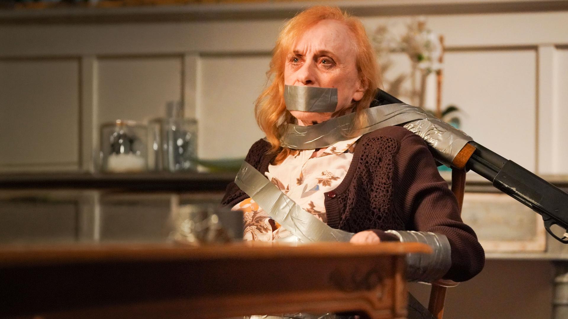 "The Rookie - Menschenjagd": Sallie (Jeannie Epper) ist mit Paketband an einen Stuhl gefesselt. Sie hat einen ängstlichen Gesichtsausdruck. Ein Gewehr ist mit Tape an der Stuhllehne befestigt und auf Sallies Kopf gerichtet.