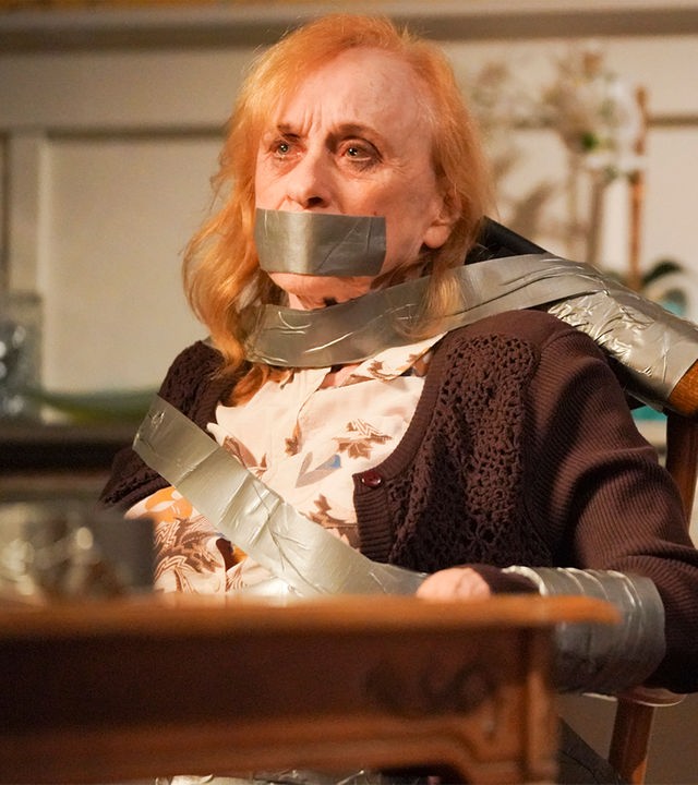 "The Rookie - Menschenjagd": Sallie (Jeannie Epper) ist mit Paketband an einen Stuhl gefesselt. Sie hat einen ängstlichen Gesichtsausdruck. Ein Gewehr ist mit Tape an der Stuhllehne befestigt und auf Sallies Kopf gerichtet.
