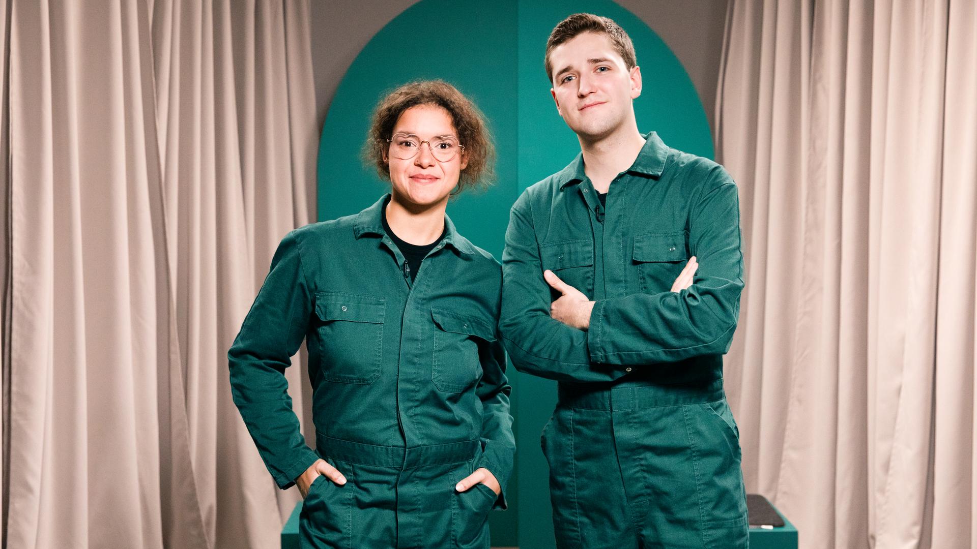 Die Gäste der Sendung, ein Mann und eine Frau, stehen im grünen Overall nebeneinander.