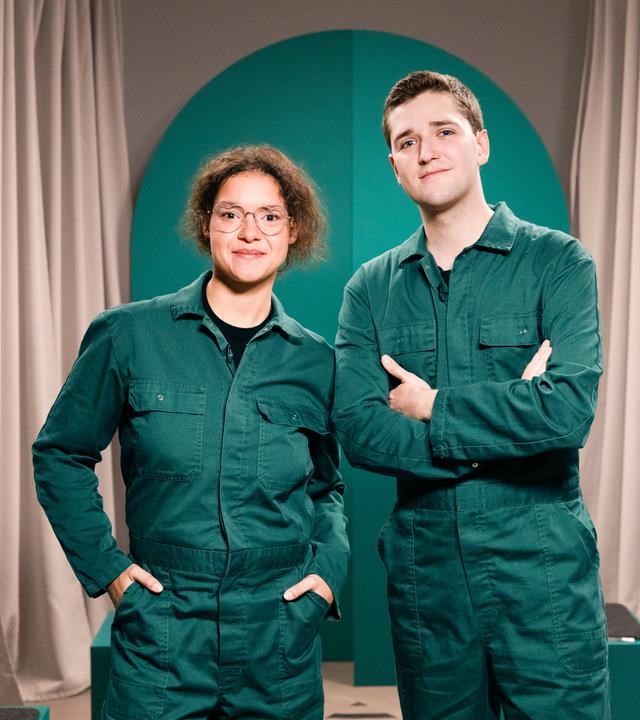 Die Gäste der Sendung, ein Mann und eine Frau, stehen im grünen Overall nebeneinander.