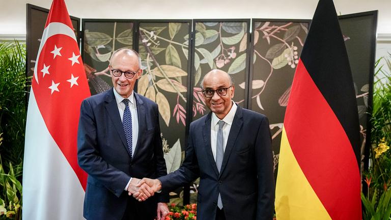 Bundeskanzler Friedrich Merz (CDU) wird vom Präsidenten der Republik Singapur, Tharman Shanmugaratnam, empfangen. Beide schauen in die Kamera und schütteln sich die Hände Im Hintergrund sieht man die Flaggen von Singapur und Deutschland.