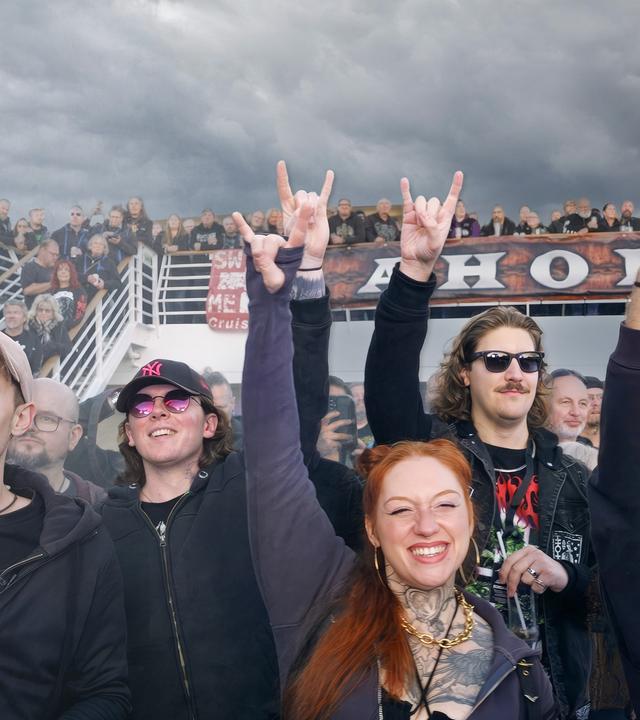 Fünf Personen strecken ihre Hände mit Zeigefinger und kleinem Finger in die Luft und jubeln einer Band zu.