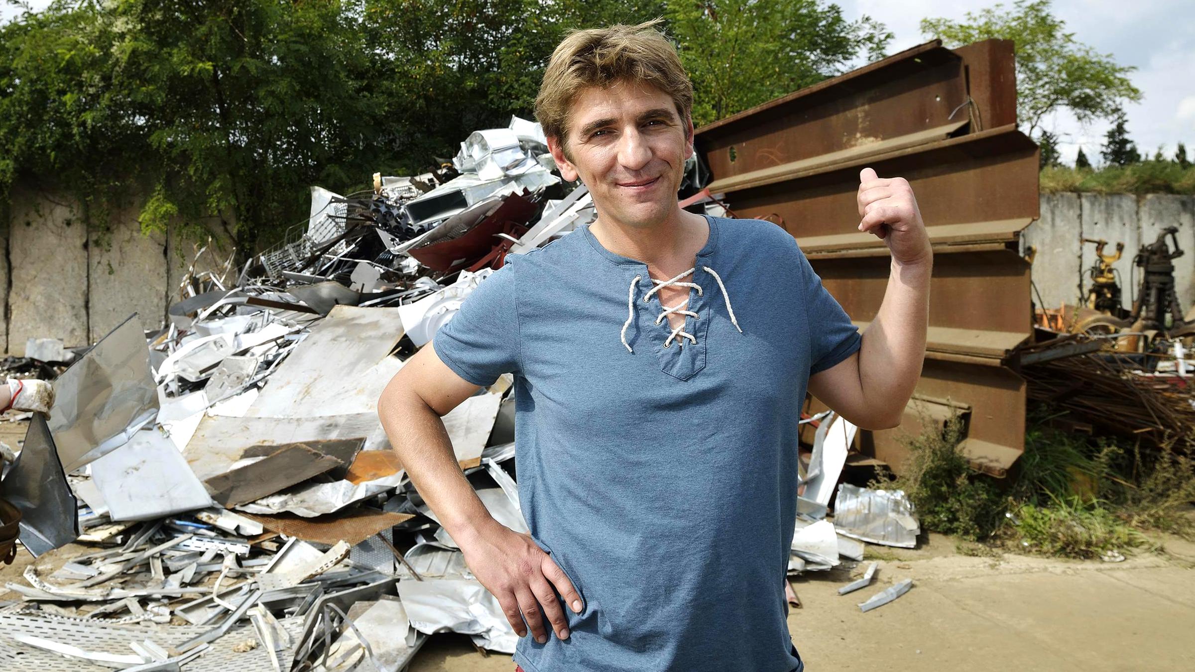 Fritz Fuchs zeigt auf einen hinter ihm liegenden Schrotthaufen. Eine Frau in lilafarbener Kleidung steht links von ihm.