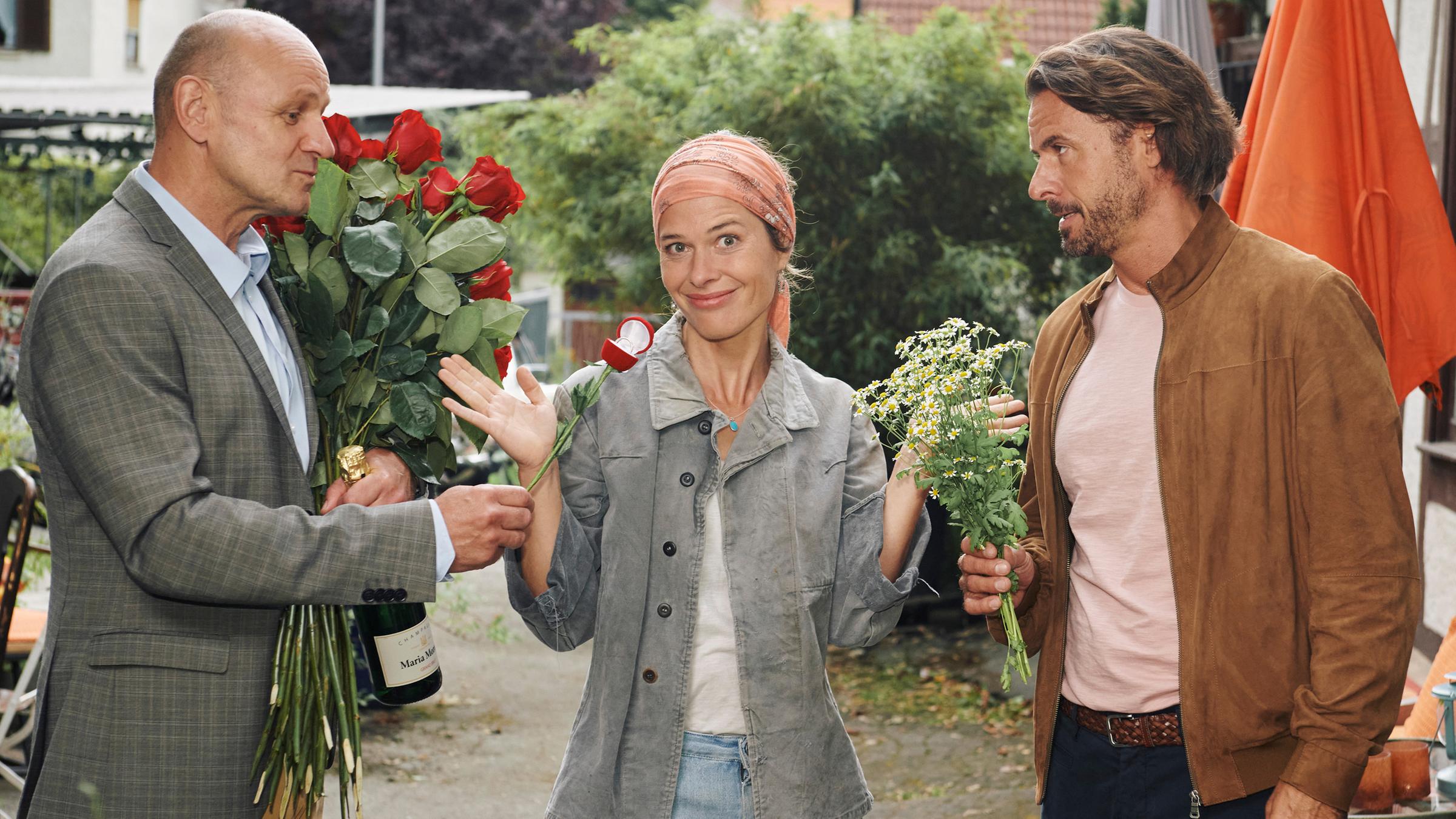 Anna (Meike Droste) steht zwischen Albert (Oliver Stokowski) und Robert (Stephan Luca) und hält ratlos die Hände in die Luft. Sie blickt gequält lächelnd in die Kamera. Albert und Robert sehen sich feindselig an, beide halten einen Blumenstrauß in den Händen, Albert hält eine Flasche Sekt und präsentiert einen Verlobungsring.