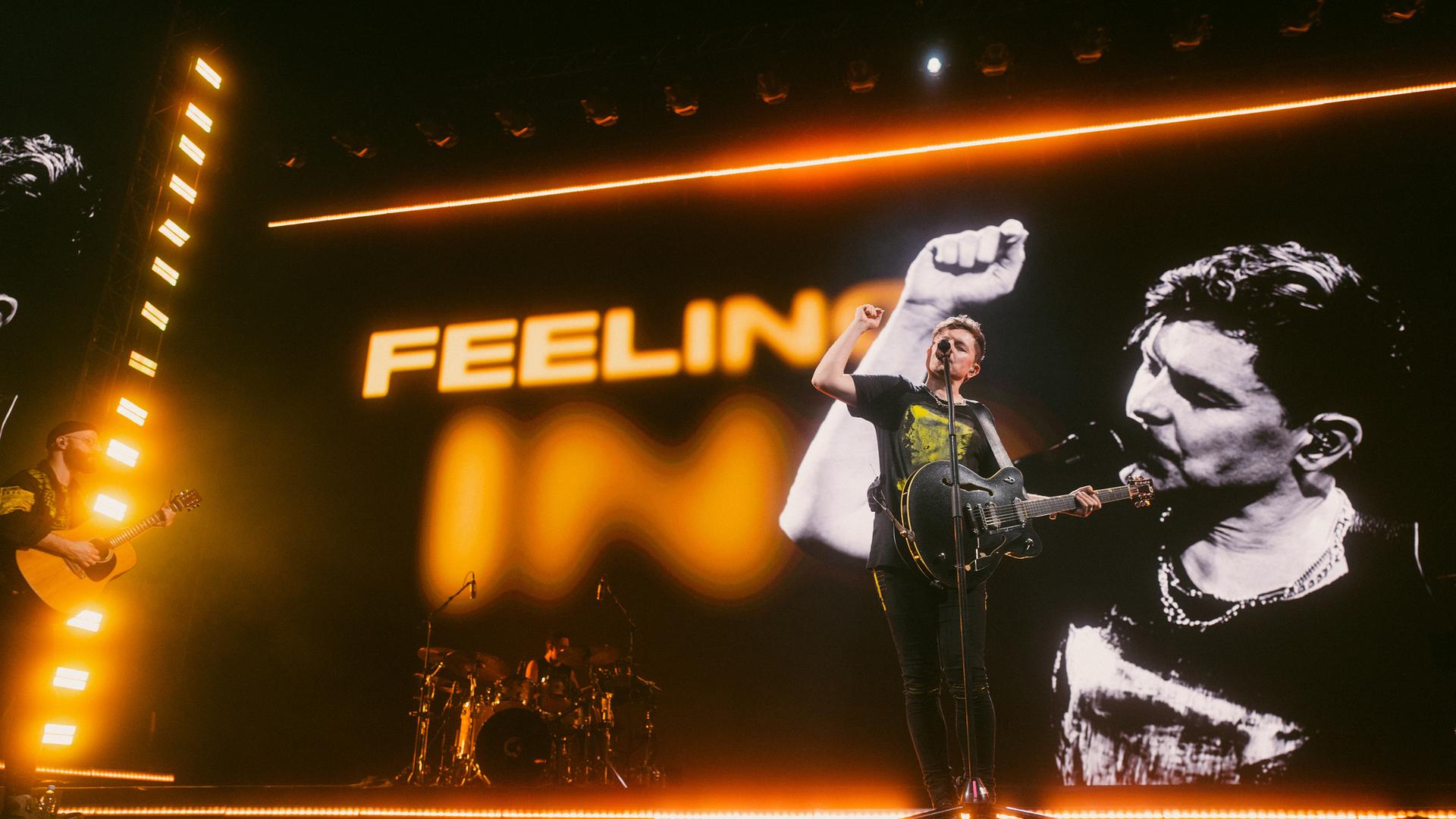 Der Sänger Michael Patrick Kelly steht mit einer Gitarre auf der Bühne, singt und hält die rechte Faust in die Luft.