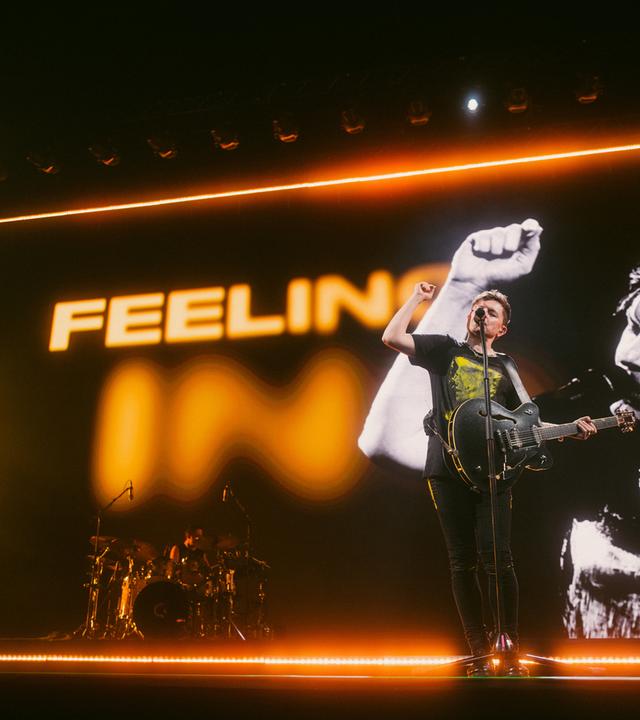 Der Sänger Michael Patrick Kelly steht mit einer Gitarre auf der Bühne, singt und hält die rechte Faust in die Luft.