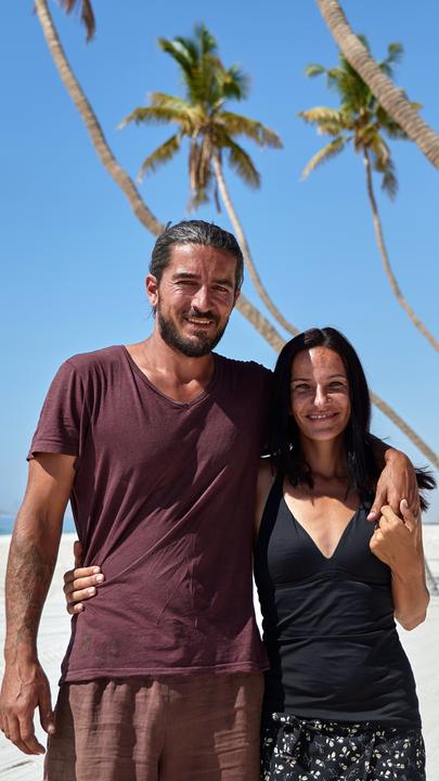 Thorben und Michi stehen Arm in Arm unter Palmen am Meer und lachen in die Kamera. Im Hintergrund ist der grüne Lkw zu sehen.