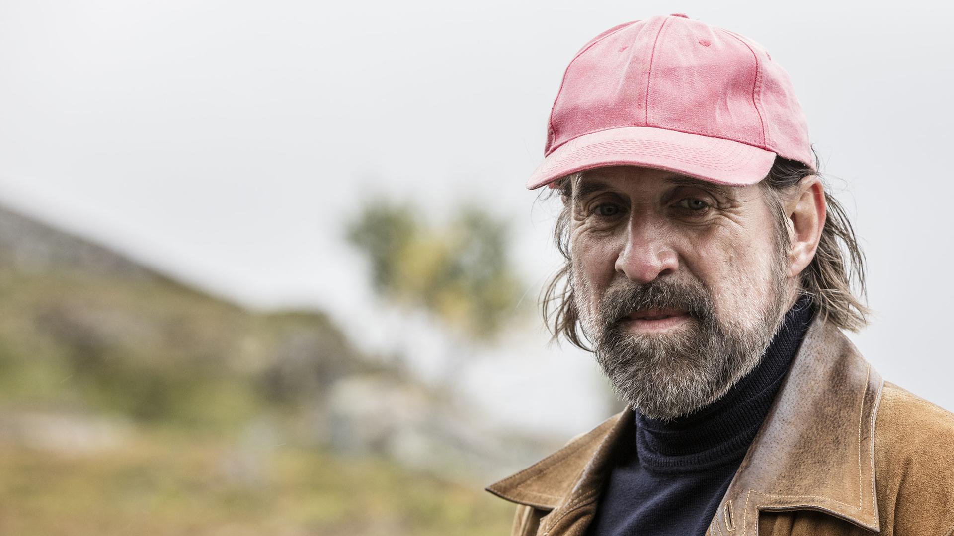 "Midnight Sun - Der fremde Tote": Rutger Burlin (Peter Stormare)  in Lederjacke und mit roter Kappe steht vor einer Berglandschaft.