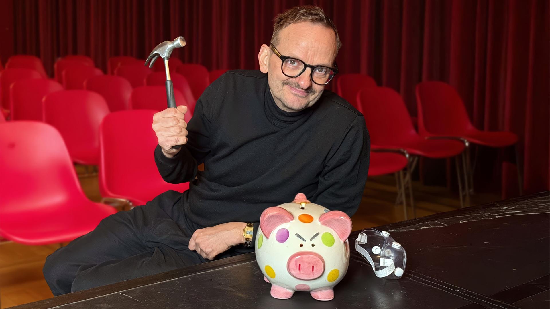 Ein Mann in schwarzer Kleidung sitzt auf einer Theaterbühne vor roten Stuhlreihen. In der Hand hält er einen Hammer, vor ihm steht ein bunt gepunktetes Sparschwein und eine Theatermaske.