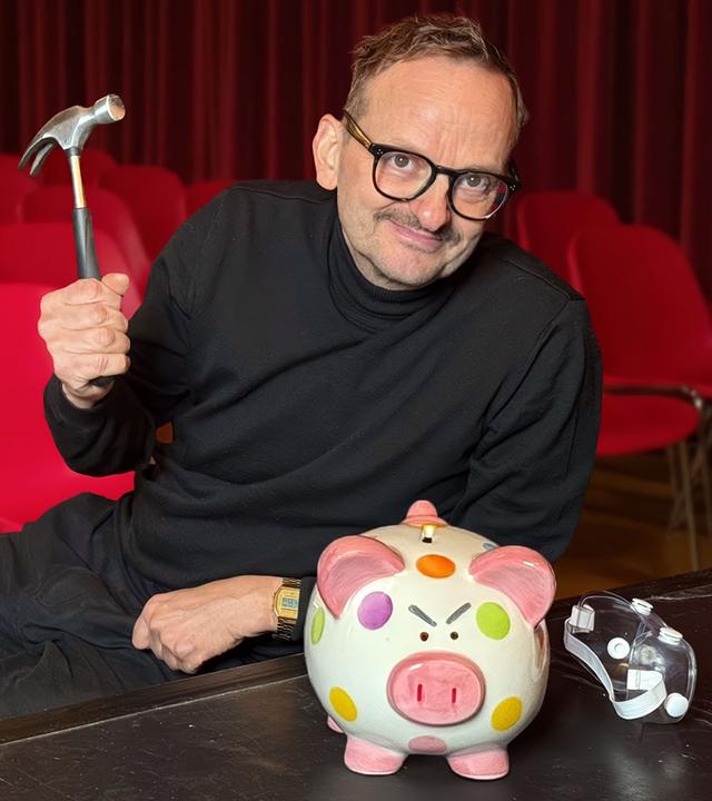 Ein Mann in schwarzer Kleidung sitzt auf einer Theaterbühne vor roten Stuhlreihen. In der Hand hält er einen Hammer, vor ihm steht ein bunt gepunktetes Sparschwein und eine Theatermaske.