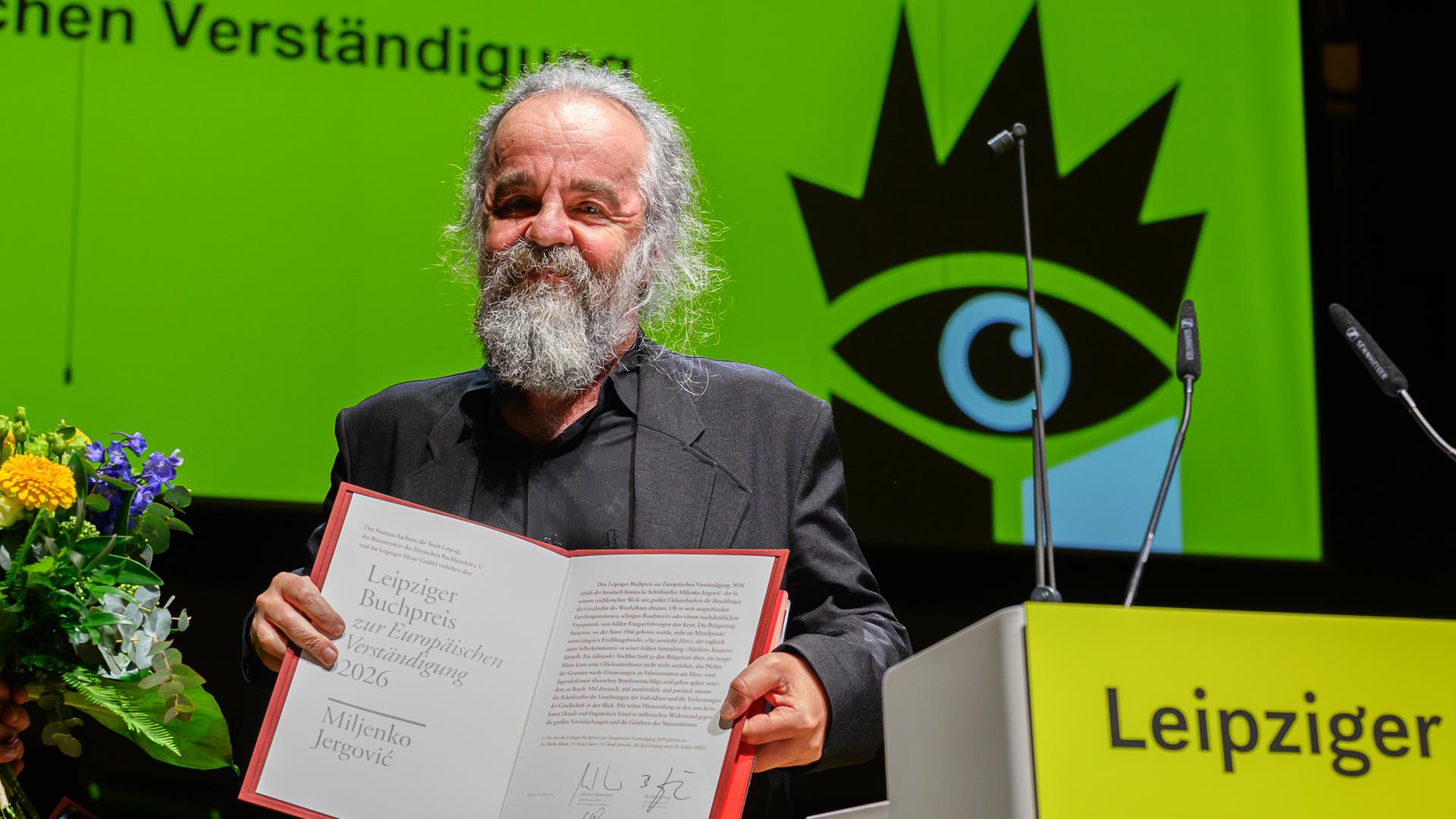 Ein Mann mit graumeliertem Bart und Haaren präsentiert auf einer Bühen eine Kladde mit einer Urkunde. Hinter ihm ist ein grünes Plakat der Leipziger Buchmesse zu sehen.