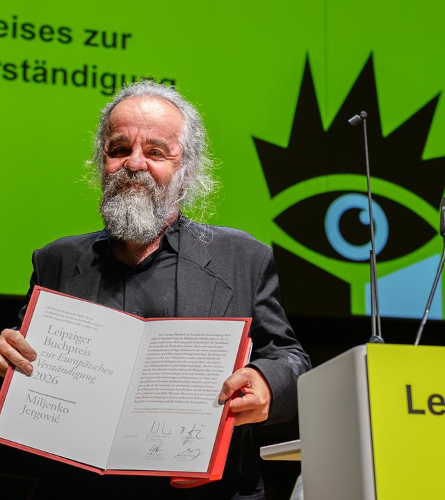 Ein Mann mit graumeliertem Bart und Haaren präsentiert auf einer Bühen eine Kladde mit einer Urkunde. Hinter ihm ist ein grünes Plakat der Leipziger Buchmesse zu sehen.