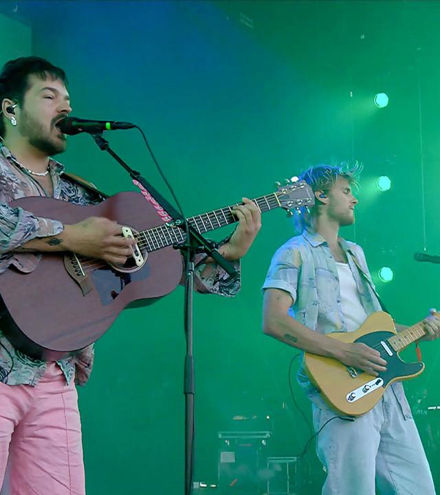 Das Bild zeigt zwei Musiker auf einer Bühne während eines Konzerts. Sie gehören zum Musikduo Milky Chance. Der linke Musiker spielt eine akustische Gitarre und trägt ein buntes Hemd sowie rosa Hosen. Er hat kurze, dunkelbraune Haare und steht in einem Mikrofon, um zu singen.   Der rechte Musiker spielt eine elektrische Gitarre und trägt ein helles, kurzärmliges Hemd über einem weißen T-Shirt. Seine Haare sind blond und leicht zerzaust. Im Hintergrund ist eine grüne Lichtinstallation zu sehen, die die Bühne beleuchtet. Die Bühne scheint Teil eines Festivals zu sein, umgeben von einem musikalischen Equipment wie Tasteninstrumenten.   Die gesamte Atmosphäre ist lebhaft und energetisch, was auf die Performancestimmung des Konzerts hinweist.
