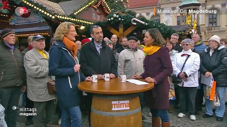 ZDF-Mittagsmazin Vor Ort in Magdeburg (12.12.2019): Annegret Oster (l.) und Jana Pareigis (r.) stehen mit einem Gesprächspartner an einem Tisch auf dem Magdeburger Weihnachtsmarkt. Im Hintergrund Passanten und weihnachtlich geschmückte Verkaufsstände.