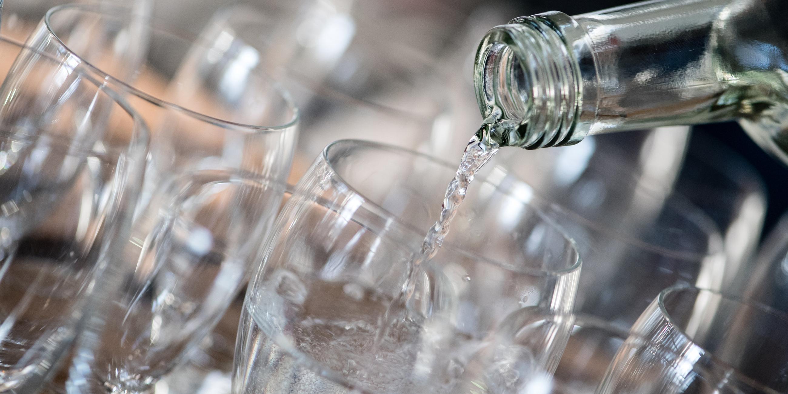 Mehrere Gläser stehen nebeneinander auf einem Tablet. Aus einer Glasflasche wird Wasser in ein Glas gegossen.