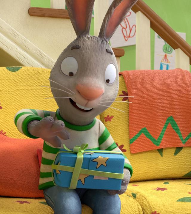 Pip sitzt auf einem Sofa und beginnt freudig, ein in Sternenpapier eingewickeltes Geschenk auszupacken.