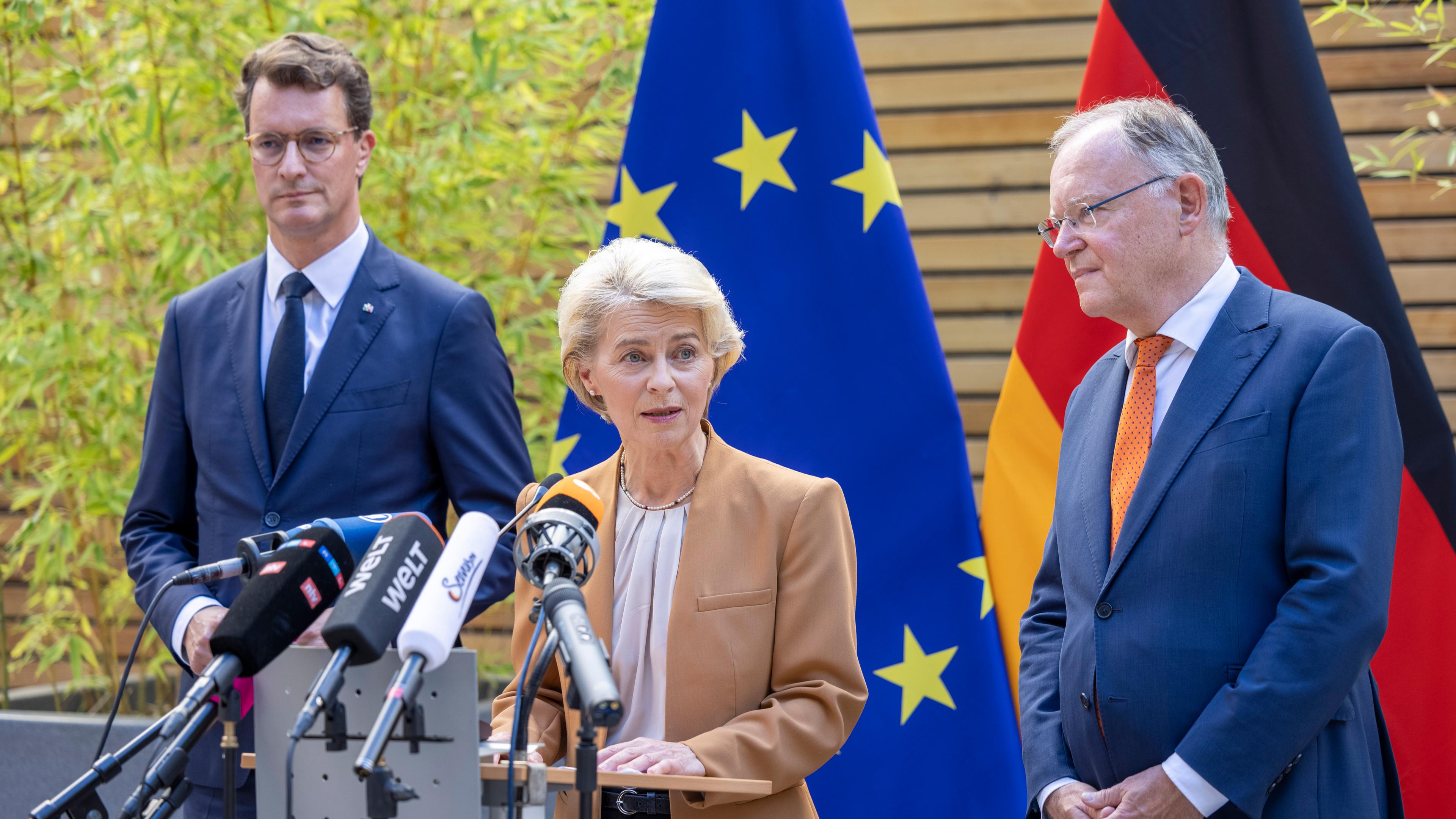 Ursula von der Leyen, Hendrik Wüst und Stephan Weil stehemn an einem Rednerpult vor einer Deutschland und einer EU-Flagge.