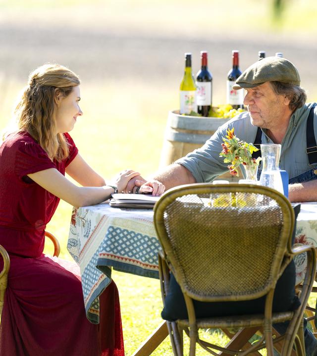 Minna (Meriel Hinsching) hat einen großen Traum: Sie möchte im hohen Norden auf dem Landgut ihres Großvaters Christer (Uwe Rohde) ein Weingut etablieren. "Inga Lindström: Minnas Traum": Minna (Meriel Hinsching) sitzt mit Christer (Uwe Rohde) an einem Tisch auf einer Wiese. Hinter ihnen stehen auf einem Tisch verschiedene Weinflaschen.