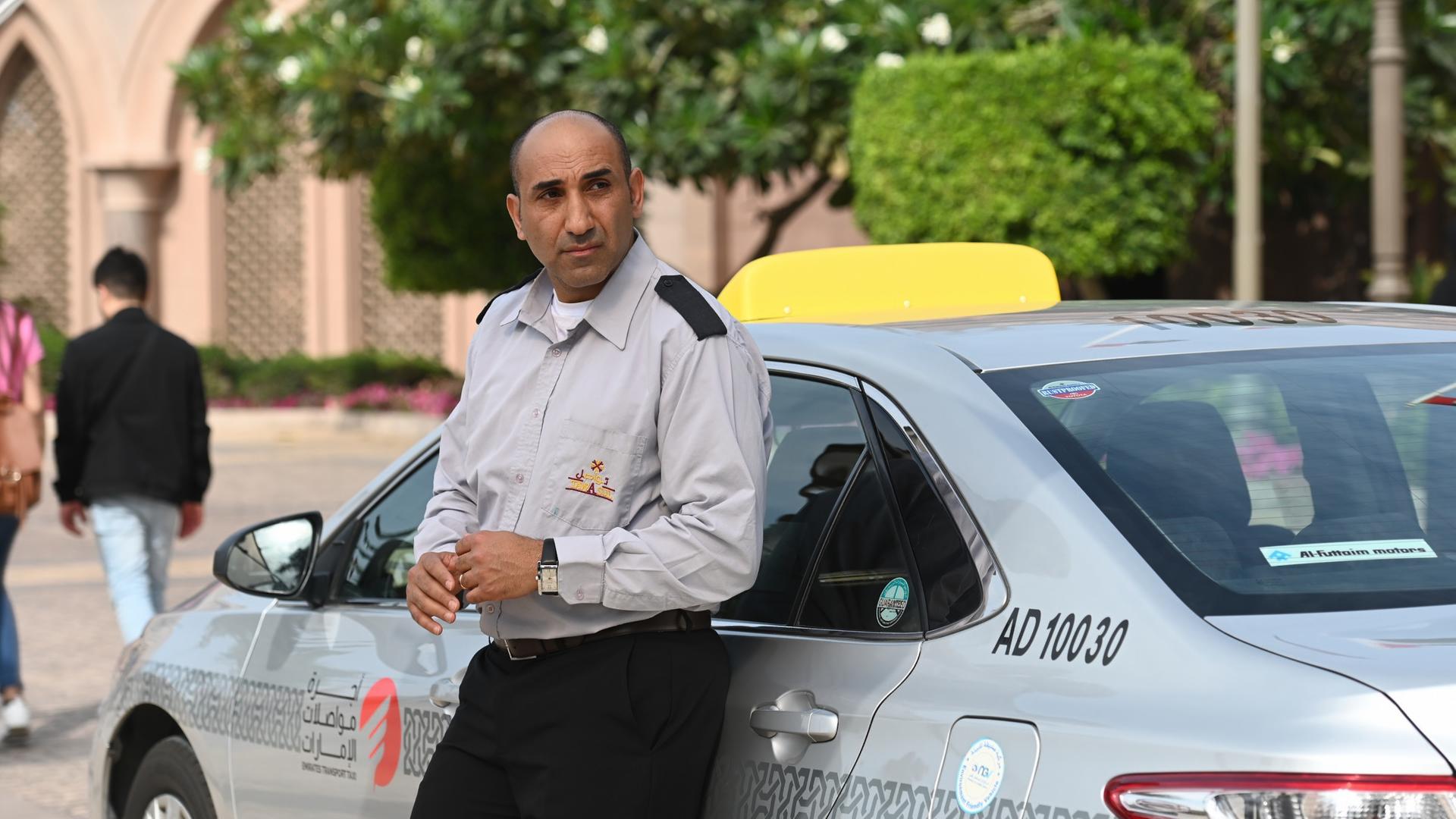 "Mirage - Gefährliche Lügen": Taxifahrer Bassem (Chadi Alhelou) lehnt in seiner Uniform an seinem Taxi und sieht zur Seite.