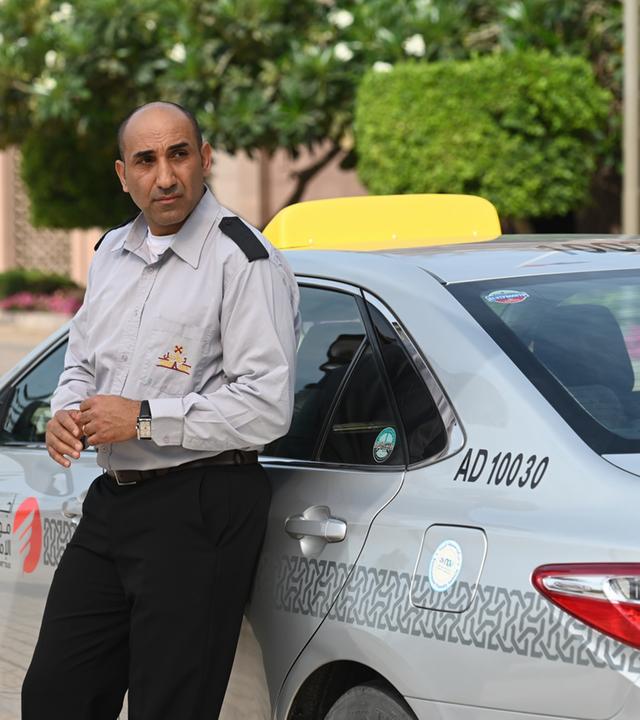 "Mirage - Gefährliche Lügen": Taxifahrer Bassem (Chadi Alhelou) lehnt in seiner Uniform an seinem Taxi und sieht zur Seite.