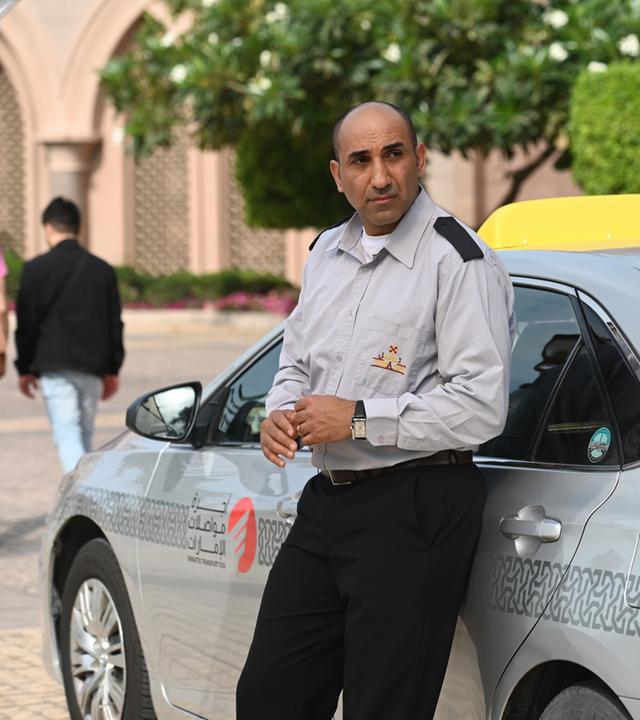 "Mirage - Gefährliche Lügen": Taxifahrer Bassem (Chadi Alhelou) lehnt in seiner Uniform an seinem Taxi und sieht zur Seite.