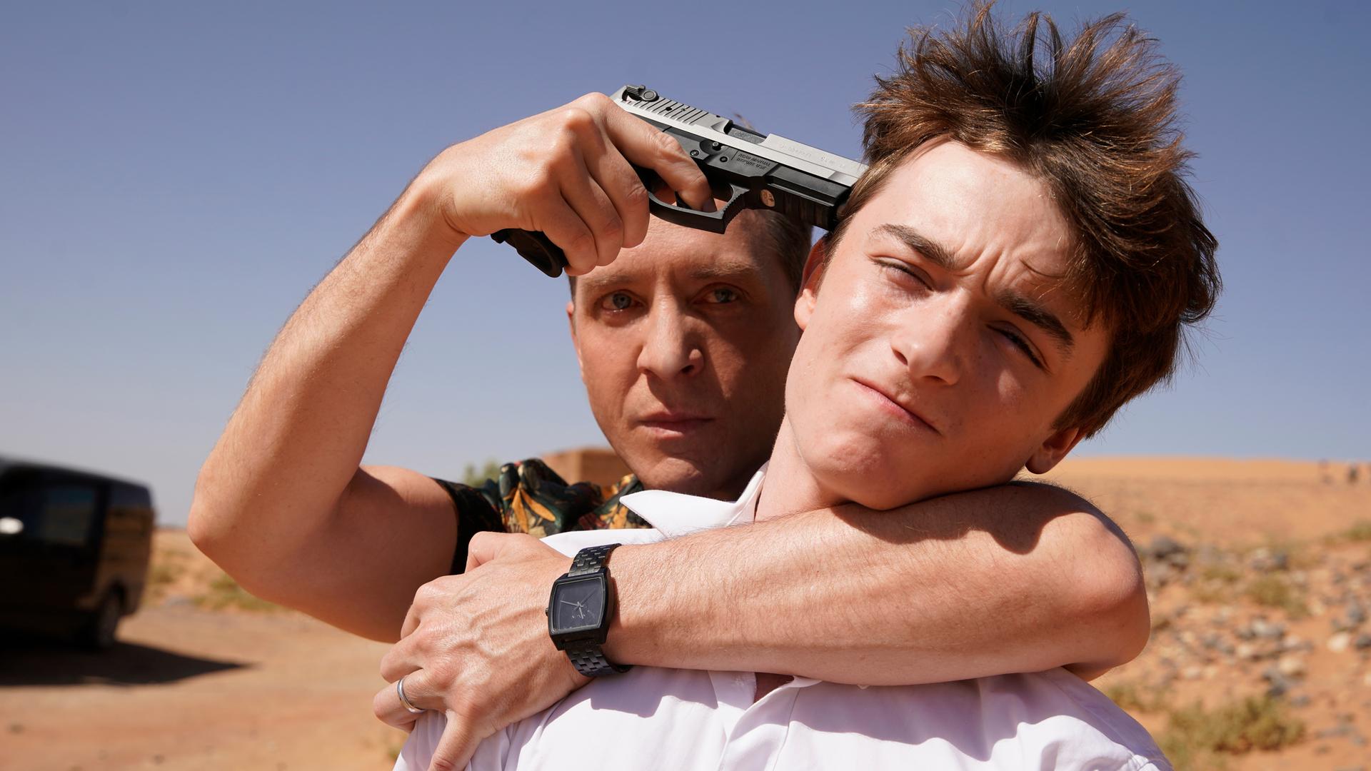 "Mirage - Gefährliche Lügen": Doug (Shawn Doyle) steht hinter Zack (Thomas Chomel). Er hat einen Arm um den Hals des Jungen gelegt und hält ihm eine Waffe an den Kopf.