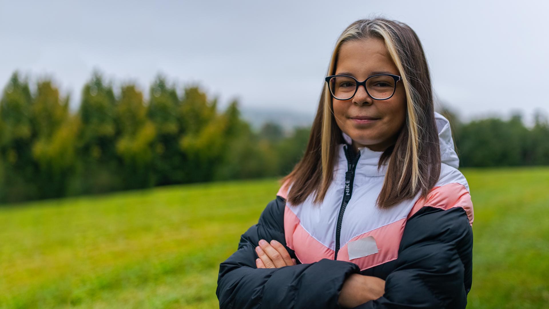 Miri steht auf einer Wiese mit verschränkten Armen. Sie sieht selbstbewusst direkt in die Kamera.