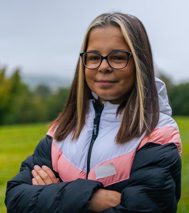 Miri steht auf einer Wiese mit verschränkten Armen. Sie sieht selbstbewusst direkt in die Kamera.