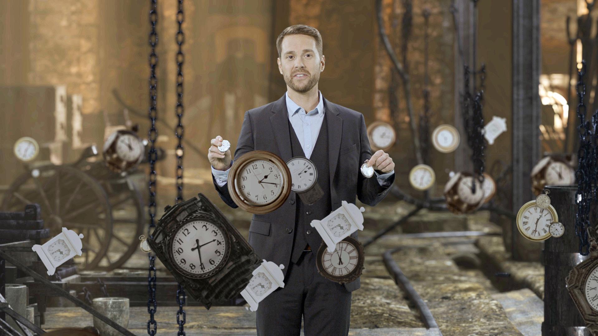 Mirko Drotschmann hält zwei Taschenuhren in den Händen und ist von Uhren umgeben.