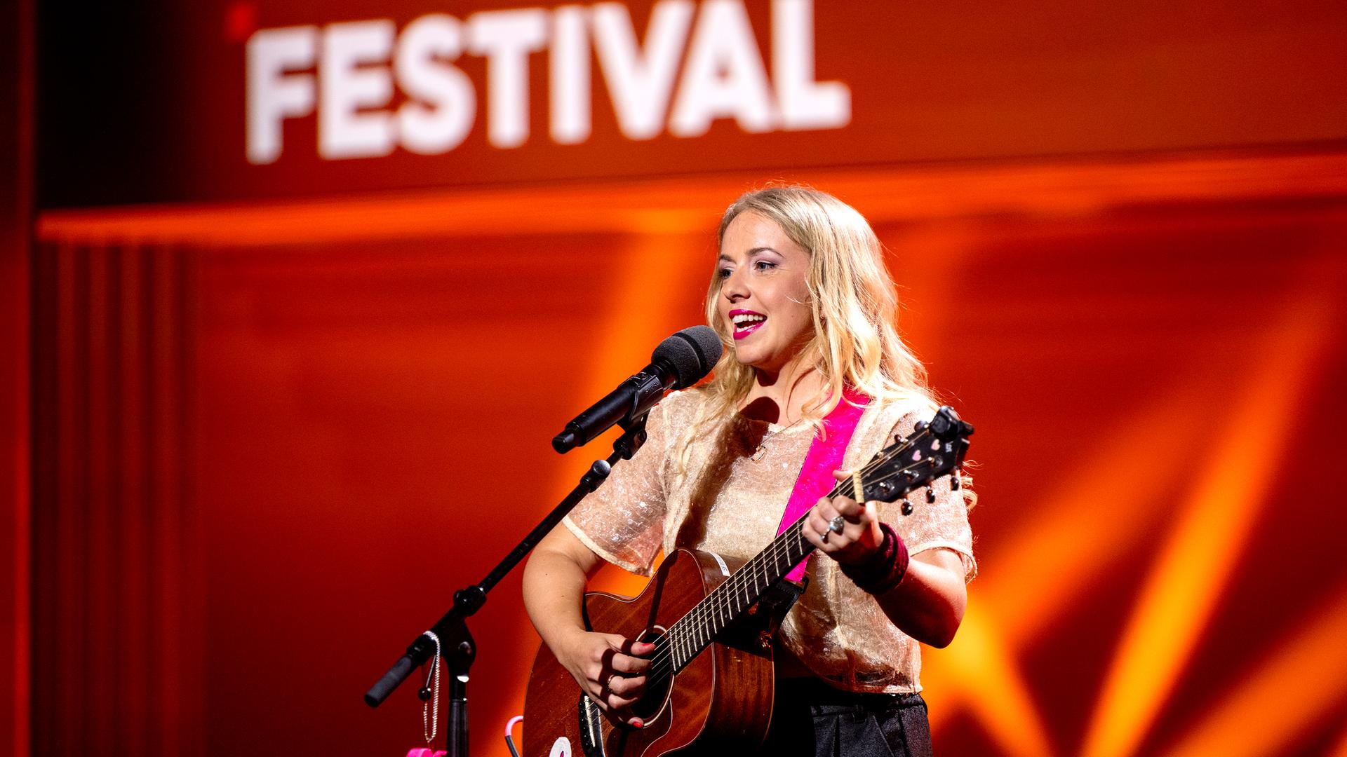 Miss Allie steht mit Gitarre in der Mitte des Bildes. Vor ihr ist ein Mikrofon. Der Hintergrund ist rot beleuchtet.