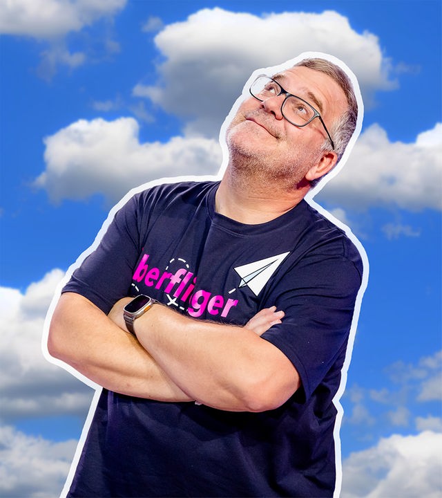Elton steht vor einem blauen Himmel mit Wolken, die Arme verschränkt. Er blickt leicht lächelnd nach oben.