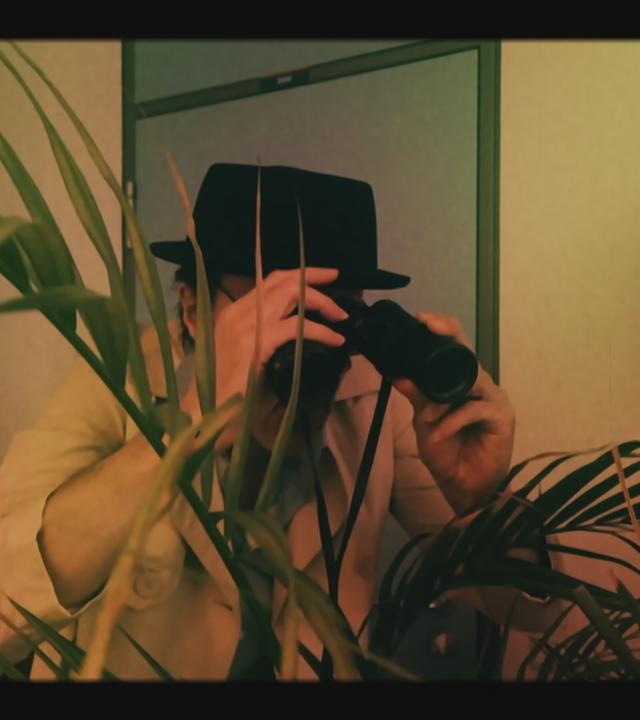 Mann mit Hut un Fernglas spioniert durch Pflanze