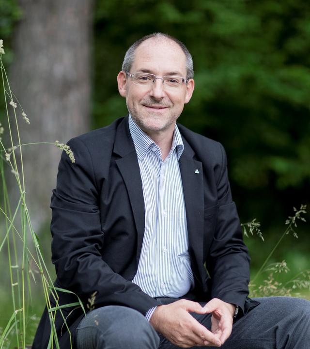 Prof. Christian Rutishauser sitzt im grünen im Hintergrund ist unscharf ein Baum zu sehen. 