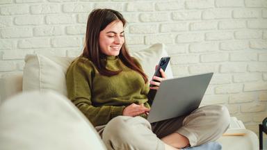Eine junge Frau sitzt im Schneidersitz auf der Couch. Sie schaut lächelnd ins Handy und hat ein offenes Laptop im Schoß.