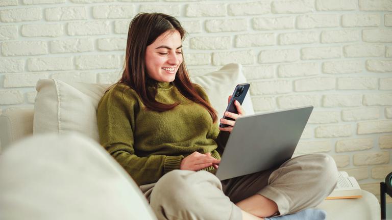 Eine junge Frau sitzt im Schneidersitz auf der Couch. Sie schaut lächelnd ins Handy und hat ein offenes Laptop im Schoß.