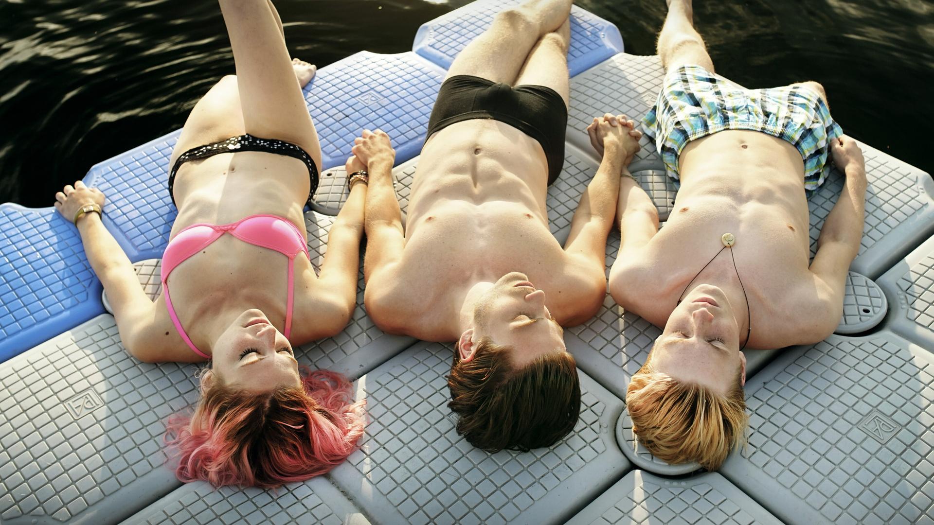 Das Bild zeigt drei Jugendliche, die auf einer schwimmenden Plattform im Wasser liegen. Von links nach rechts ist ein Mädchen mit kurzen, pinken Haaren und einem Bikinioberteil in hellrosa zu sehen, sie hat ihre Augen geschlossen und liegt entspannt. Neben ihr befindet sich ein Junge mit schulterlangen, dunklen Haaren, der nackt von der Taille nach oben zu sehen ist, seine Körperhaltung ist locker und er hält die Hand des Mädchens. Der dritte Jugendliche, ebenfalls ein Junge, hat kurze, blonde Haare und trägt eine kurze Badehose mit einem karierten Muster in Blau und Grün. Er liegt mit geschlossenen Augen und einer entspannten Haltung auf der Plattform. Der Hintergrund zeigt dunkles Wasser, und die Sonne scheint, was eine warme, sommerliche Atmosphäre schafft.