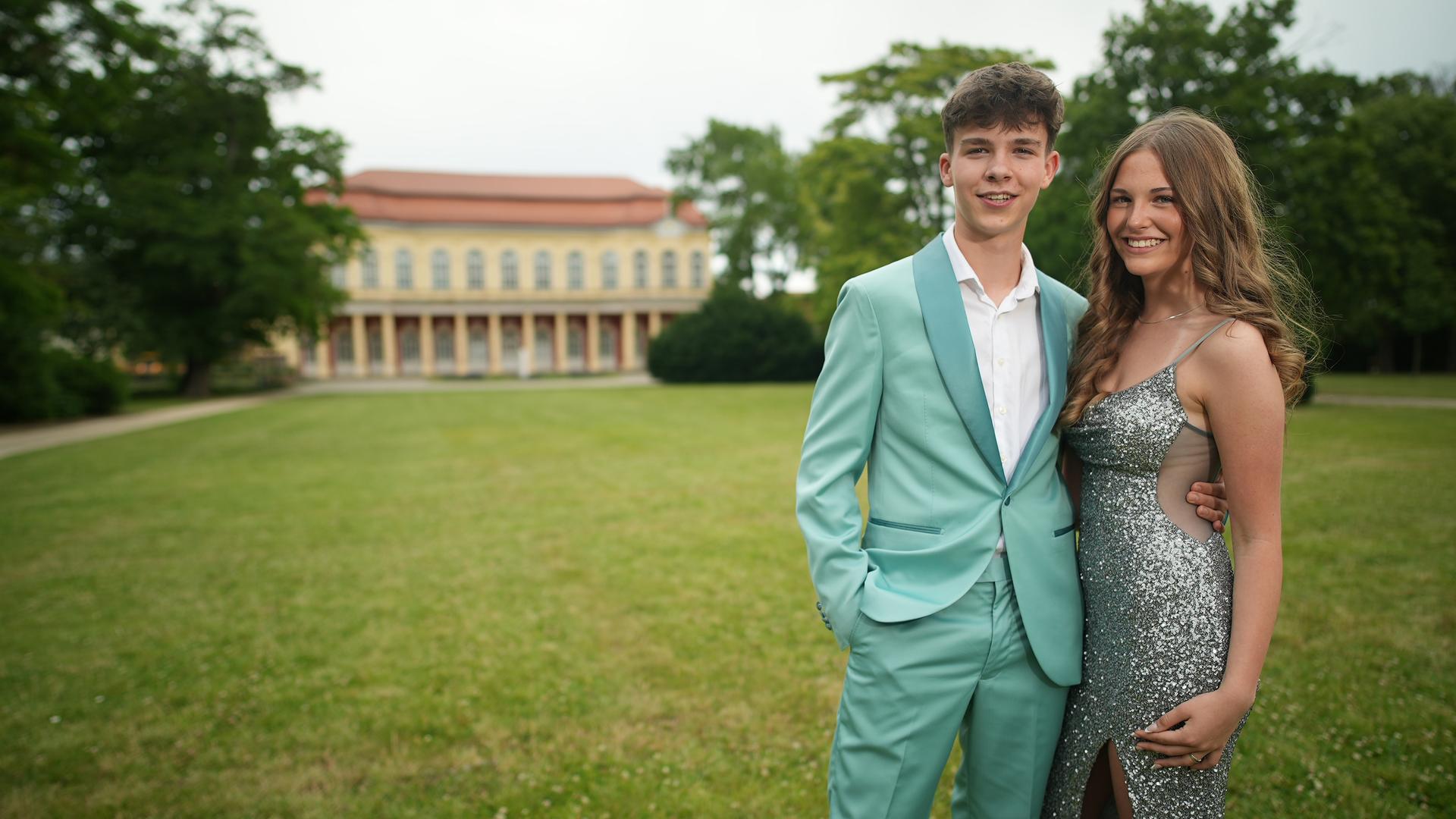 Zwei Jugendliche stehen nebeneinander. Der Junge steht links und trägt ein grünes Jackett und ein weißes Hemd, das Mädchen steht rechts neben ihm und trägt ein silbernes Paillettenkleid. 