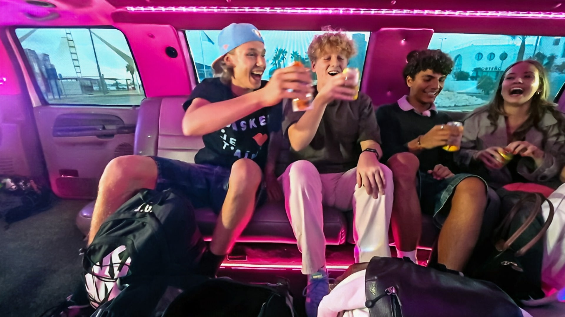 v.l.n.r. Jan, Rufus, Adrian und Lina-Marie sitzen in einer rosa ausgelichteten Limousine, lachen und stoßen mit Getränken an.