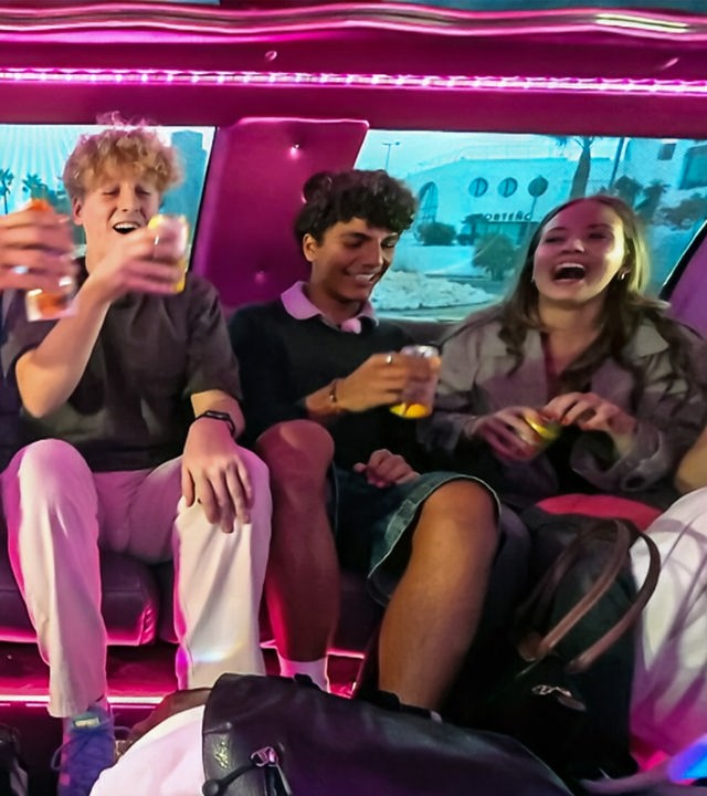 v.l.n.r. Jan, Rufus, Adrian und Lina-Marie sitzen in einer rosa ausgelichteten Limousine, lachen und stoßen mit Getränken an.