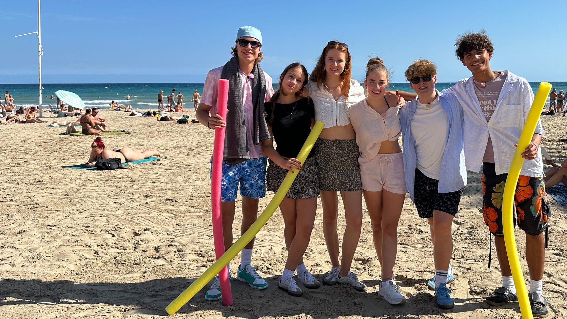v.l.n.r. Jan, Lina-Marie, Marie, Lenja, Rufus und Adrian stehen gemeinsam am Strand, legen ihre Arme umeinander und lächeln in die Kamera. Jan, Lina-Marie und Adrian haben jeweils eine Poolnudel in der Hand. Rufus und Jan tragen Sonnenbrillen.