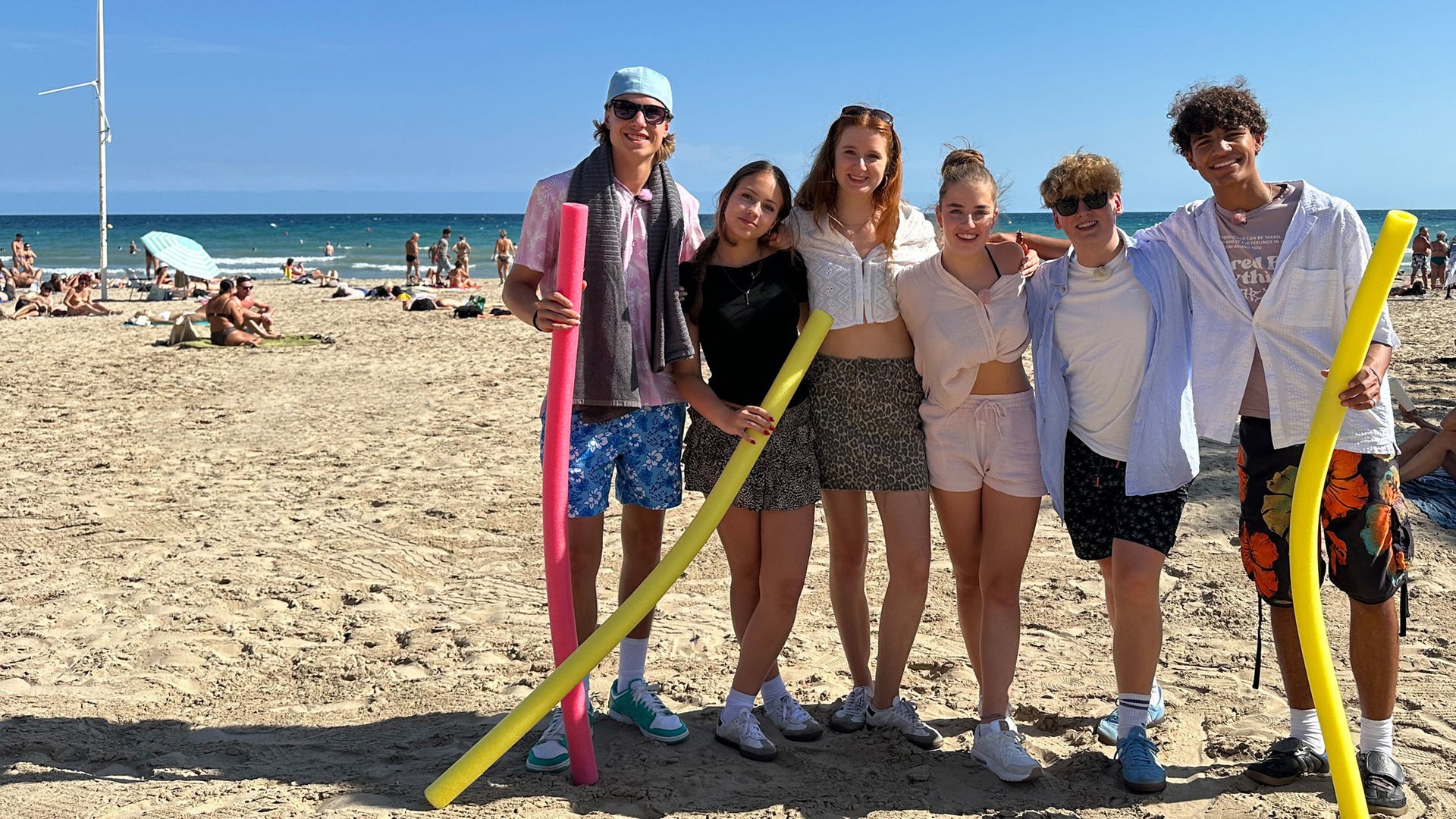 v.l.n.r. Jan, Lina-Marie, Marie, Lenja, Rufus und Adrian stehen gemeinsam am Strand, legen ihre Arme umeinander und lächeln in die Kamera. Jan, Lina-Marie und Adrian haben jeweils eine Poolnudel in der Hand. Rufus und Jan tragen Sonnenbrillen.