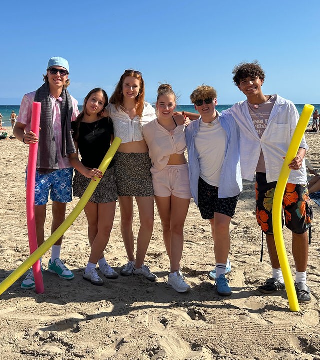 v.l.n.r. Jan, Lina-Marie, Marie, Lenja, Rufus und Adrian stehen gemeinsam am Strand, legen ihre Arme umeinander und lächeln in die Kamera. Jan, Lina-Marie und Adrian haben jeweils eine Poolnudel in der Hand. Rufus und Jan tragen Sonnenbrillen.