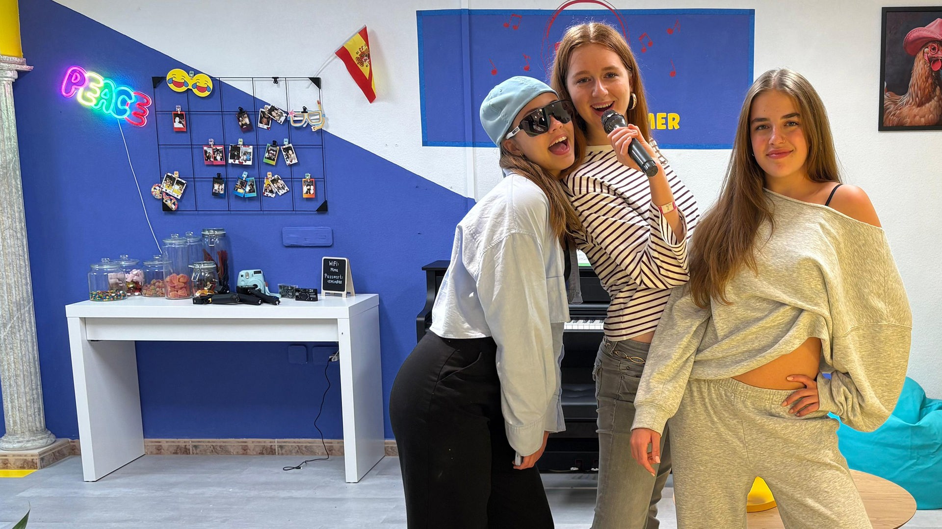 v.l.n.r. Lina-Marie, Marie und Lenja stehen nebeneinander. Marie hat ein Mikrofon in der Hand und führt es zu ihrem Mund, Lina-Marie lehnt sich dazu und öffnet ihren Mund zum Singen. Lina-Marie trägt eine Kopfbedeckung und Sonnenbrille.