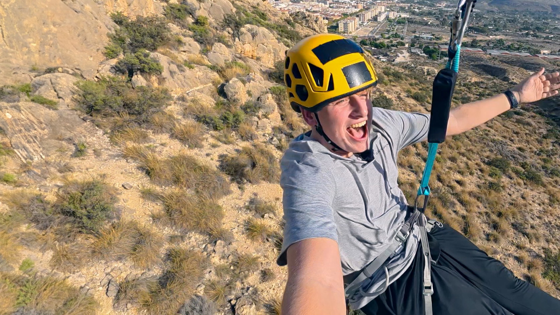 Rufus hängt an einer Zipline, hat die Kamera in der rechten Hand und schreit. Er trägt einen Helm und streckt den linken Arm aus. Im Hintergrund sind Hügellandschaften zu sehen.