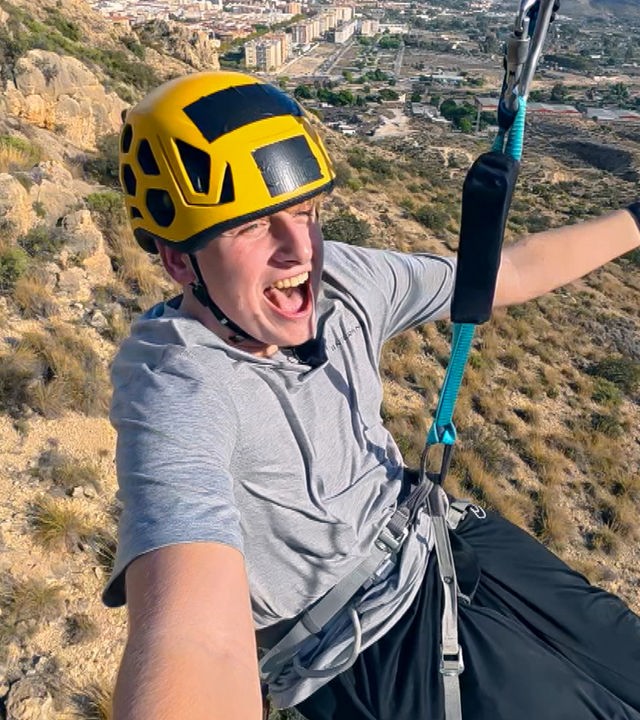 Rufus hängt an einer Zipline, hat die Kamera in der rechten Hand und schreit. Er trägt einen Helm und streckt den linken Arm aus. Im Hintergrund sind Hügellandschaften zu sehen.