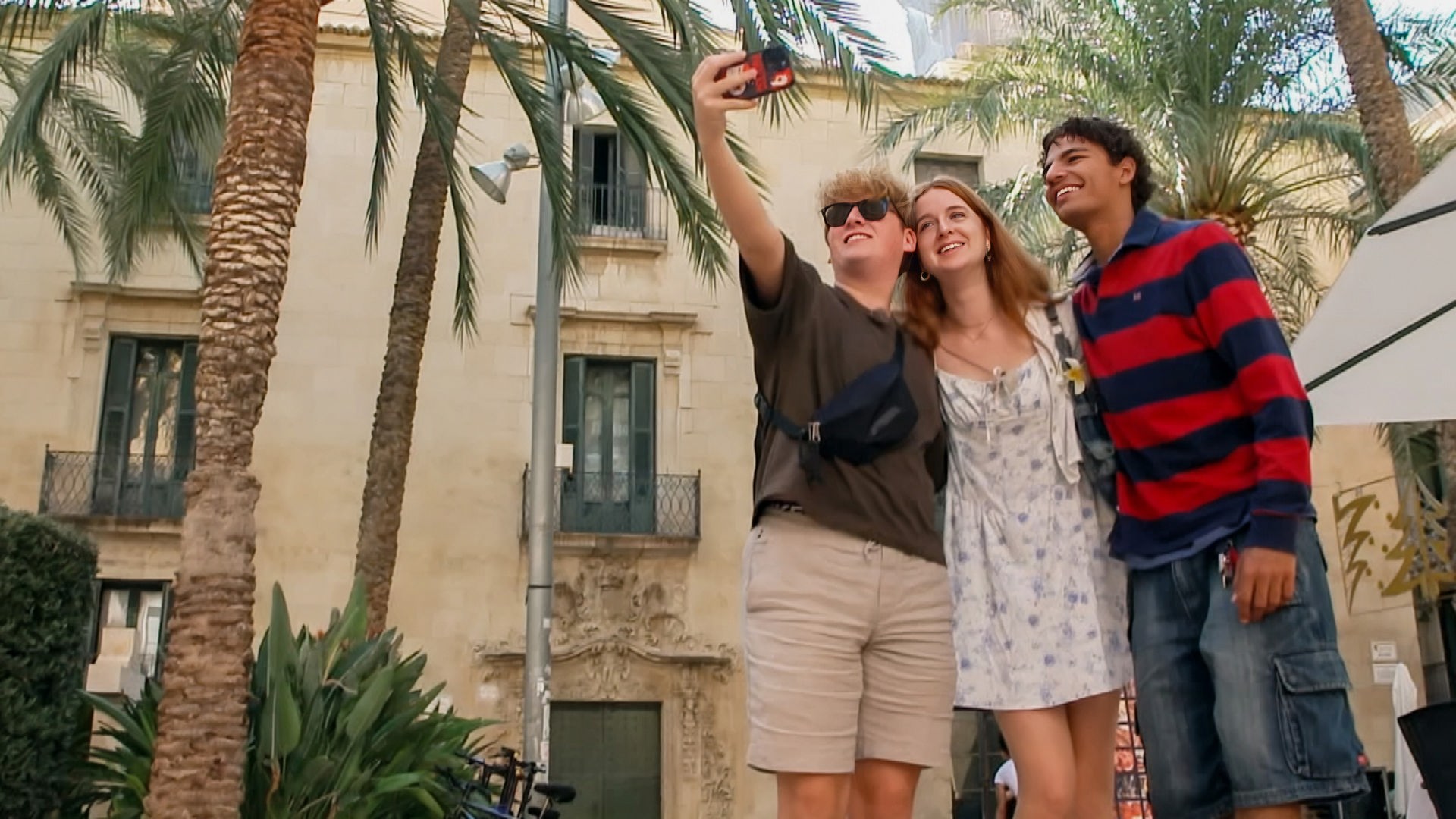 v.l.n.r. Rufus, Marie und Adrian stehen vor einem Wohnhaus neben einer Reihe Palmen und Rufus macht mit dem Handy ein Gruppenselfie.