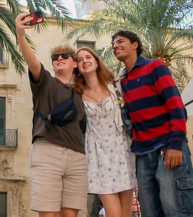 v.l.n.r. Rufus, Marie und Adrian stehen vor einem Wohnhaus neben einer Reihe Palmen und Rufus macht mit dem Handy ein Gruppenselfie.
