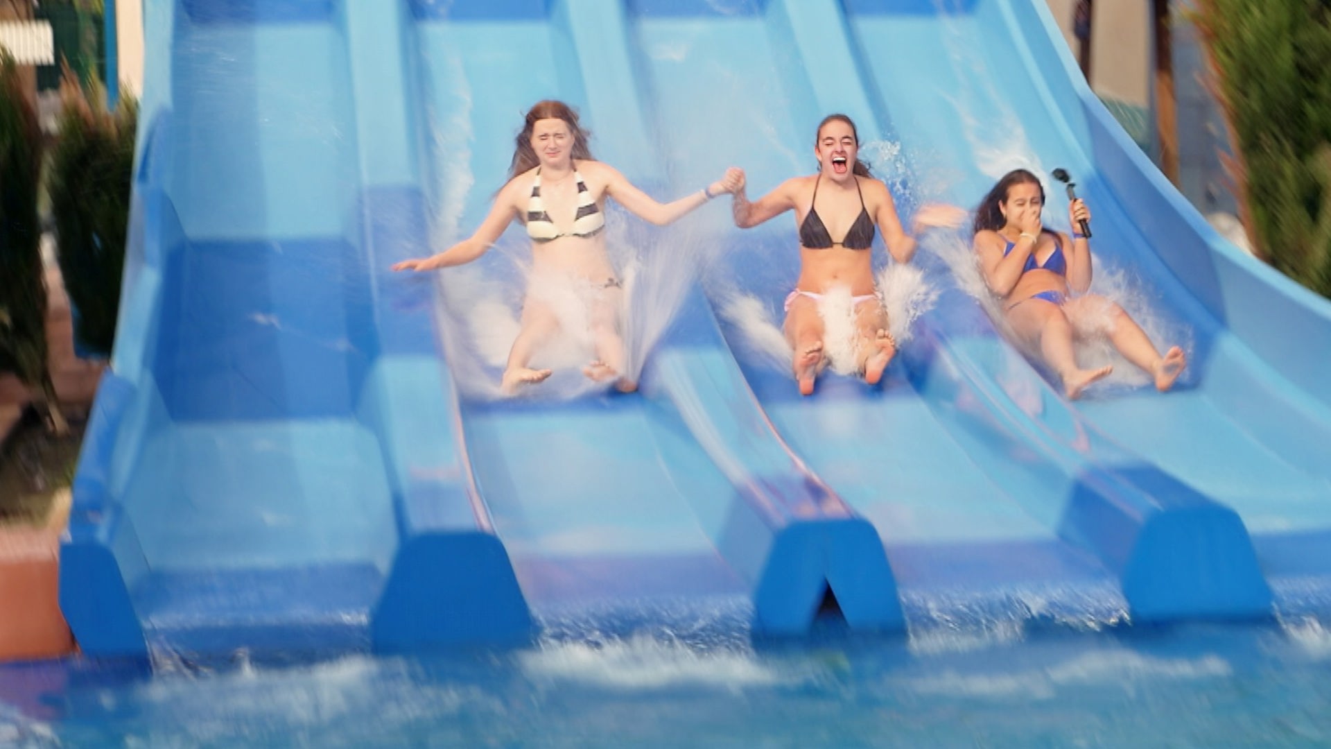v.l.n.r. Marie, Lenja und Lina-Marie rutschen auf einer blauen Reihen-Wasserrutsche nebeneinander runter. Sie halten sich gegenseitig an den Händen, Lina-Marie hat eine GoPro in der freien Hand.